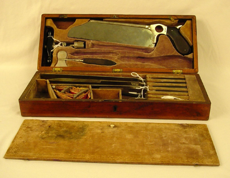 An open wooden surgical kit with metal instruments inside with wood handles. These include a bone saw, Liston knives, a tourniquet, a Hay's saw, and a trephine.